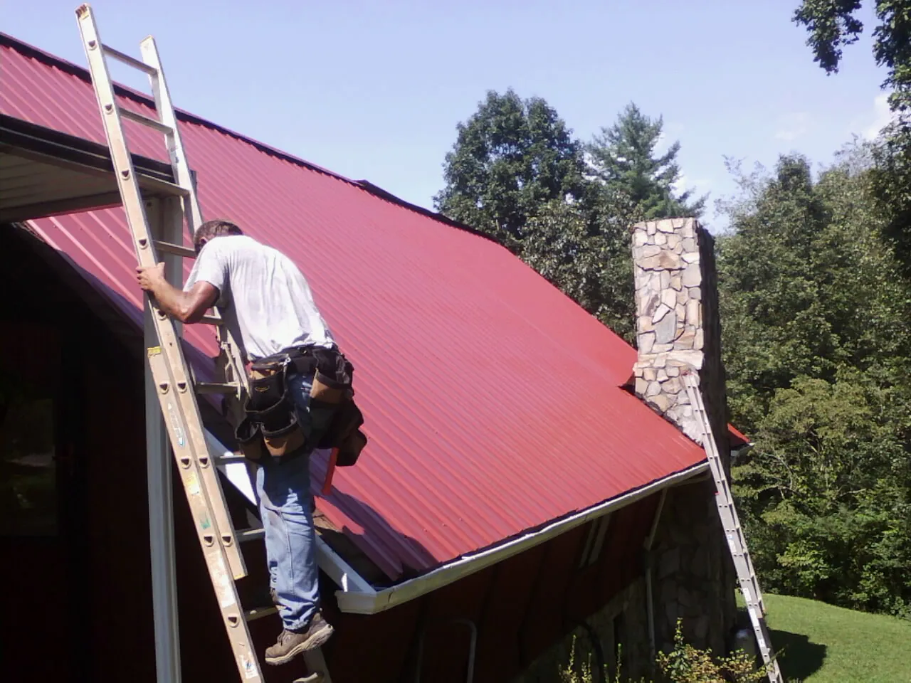Roofer on a ladder beside a red metal roof and stone chimney — Soffit & Fascia Repair by Roofing Brothers in Smithtown